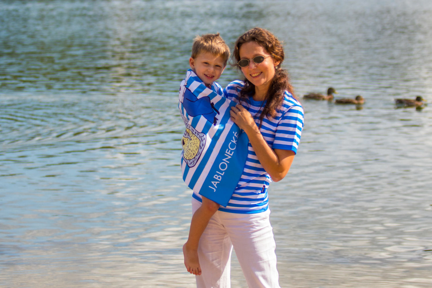 Maminka v pruhovaném tričku držící dítě v ručníku na břehu jablonecké přehrady Mšeno